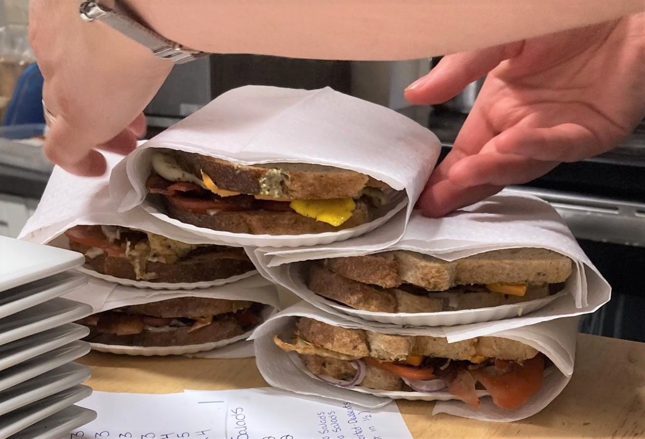 Tight shot of five plated sandwiches. A pair of hands reaches to lift one.