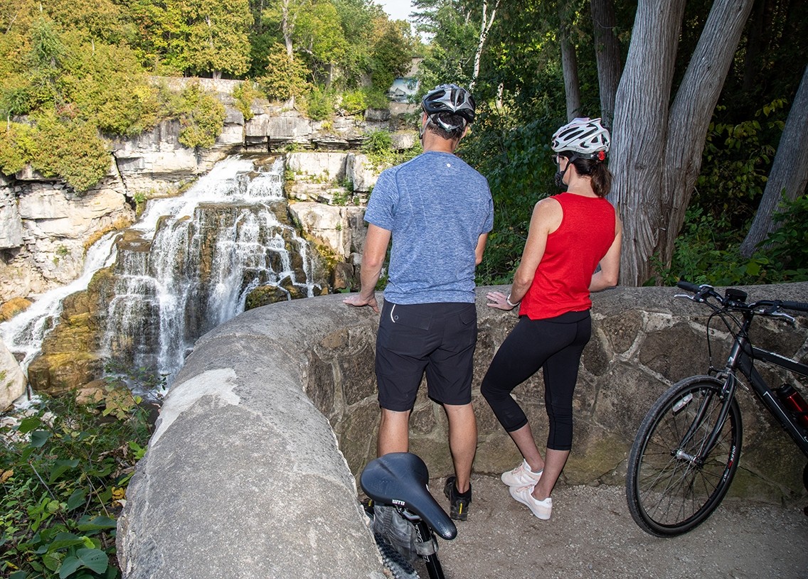 Two cyclists wearing helmets stand at a lookout observing a waterfall.
