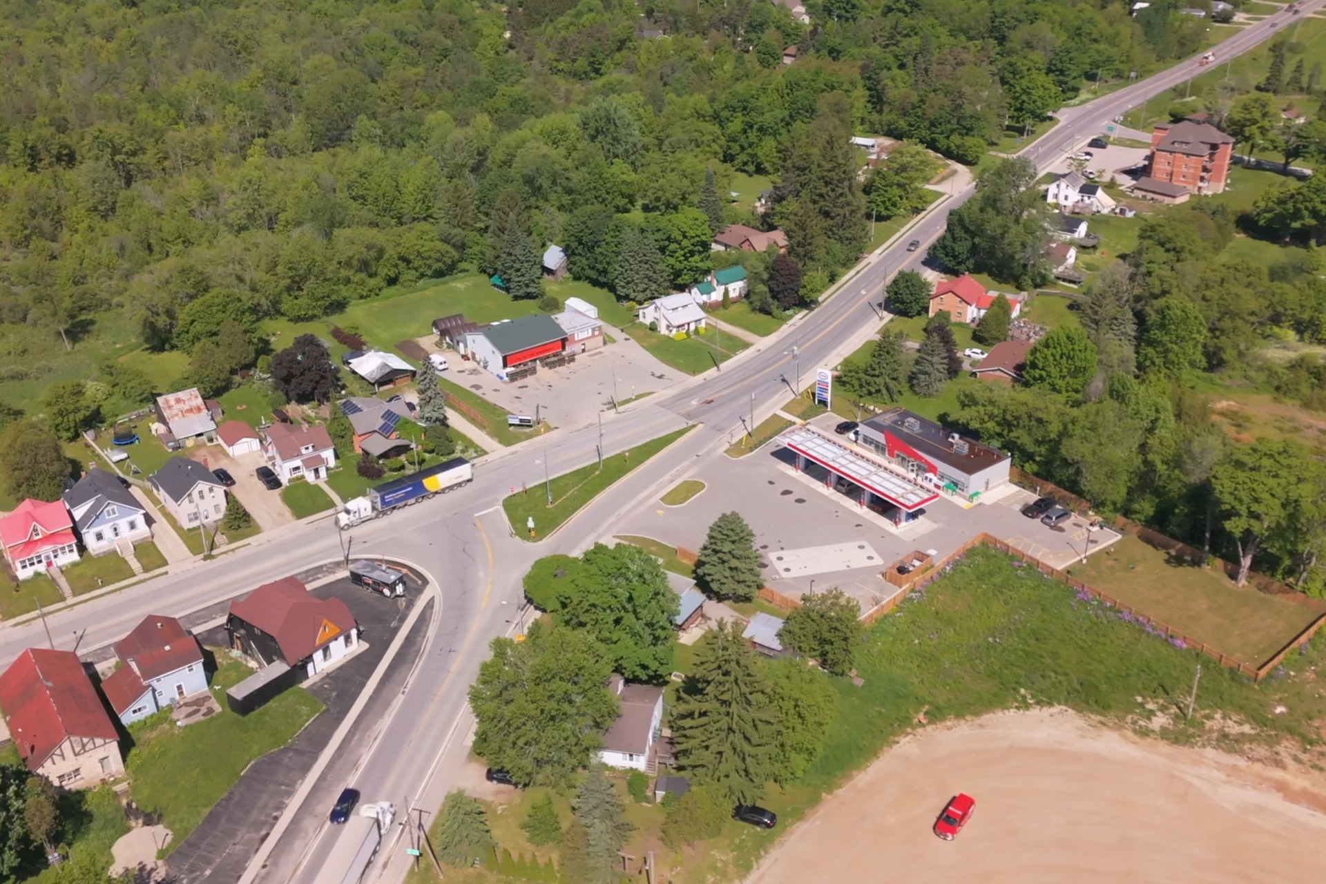 Aerial view of a small town intersection with houses, businesses, gas station, forested areas, and connecting roads.