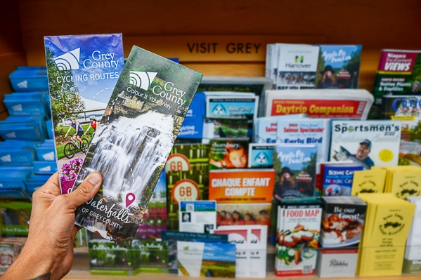 A hand holds two maps in front of a rack of brochures and magazines.