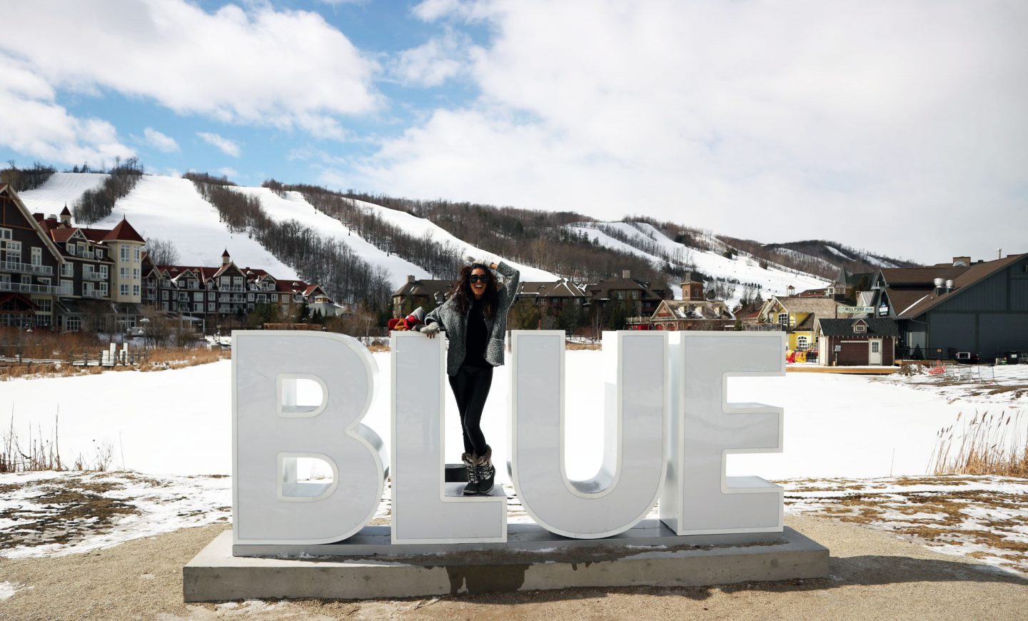 On a bright snowy day, a person stands smiling on a large sign that reads "BLUE".
