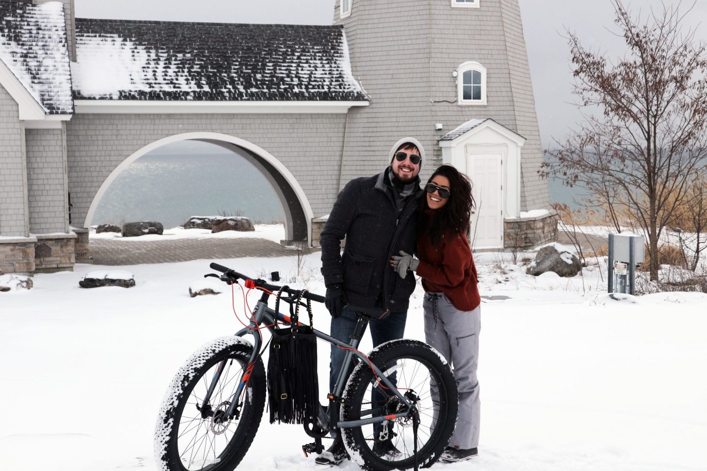 Two people stand beside a bicycle with large snow-covered tires on a winter day.