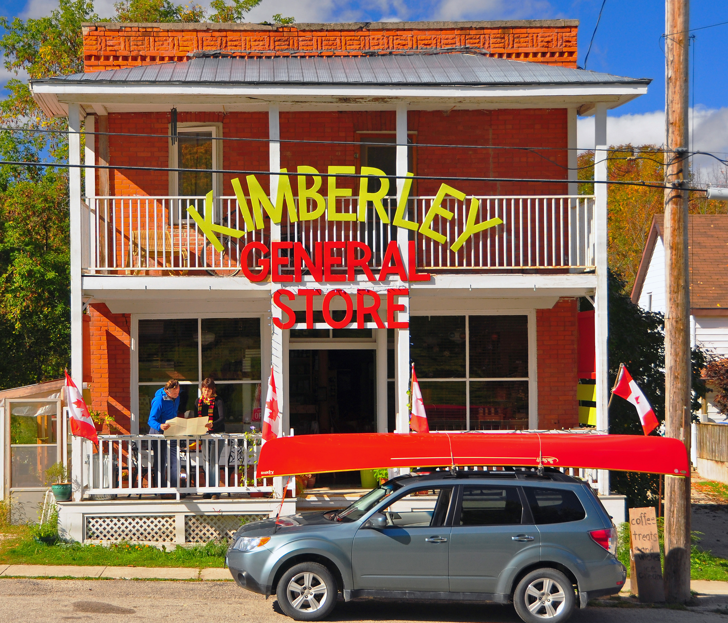 Brick general store with a balcony and a red canoe on a parked car out front.