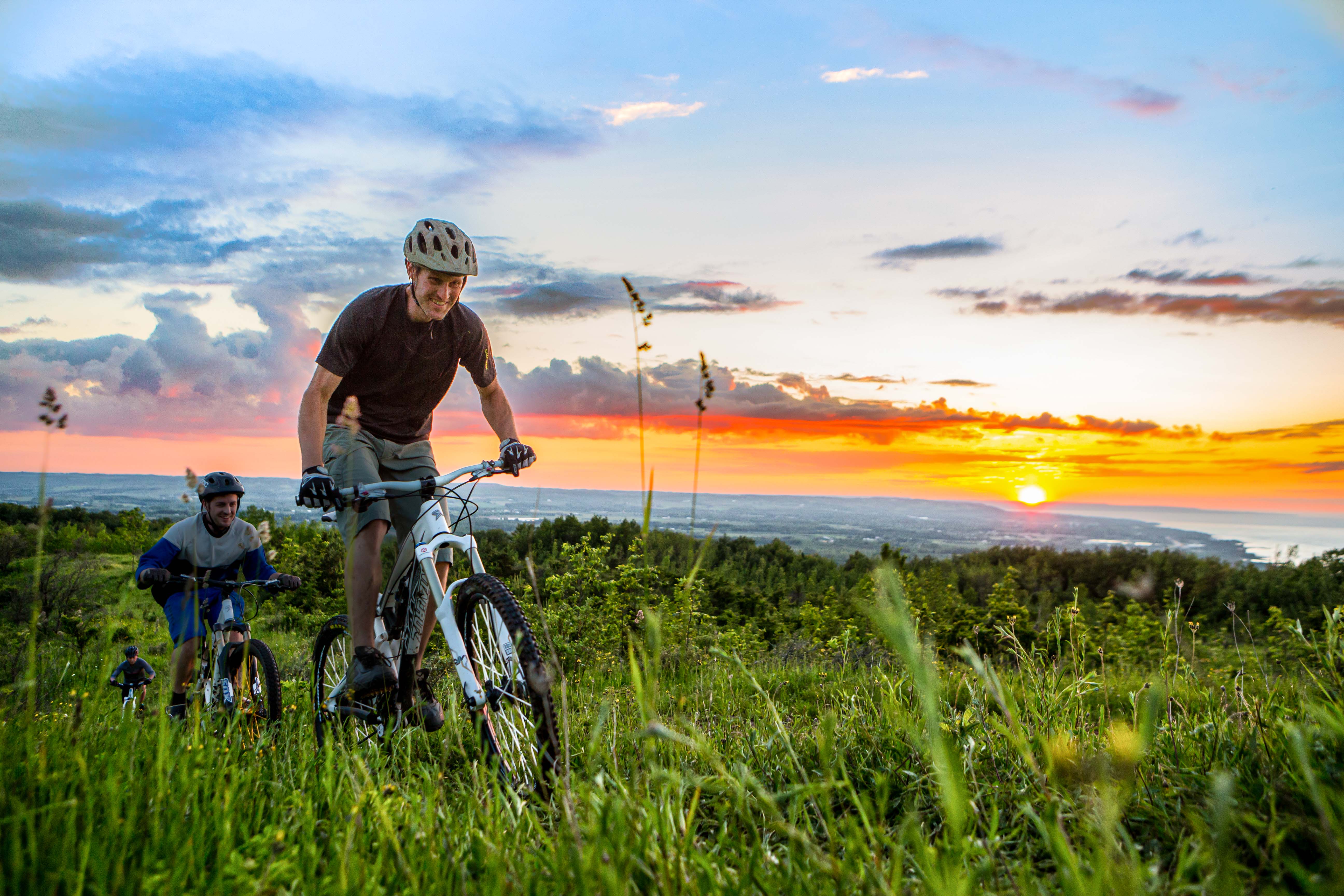 Two cyclists ride uphill through tall grass at sunset overlooking distant water and rolling hills.