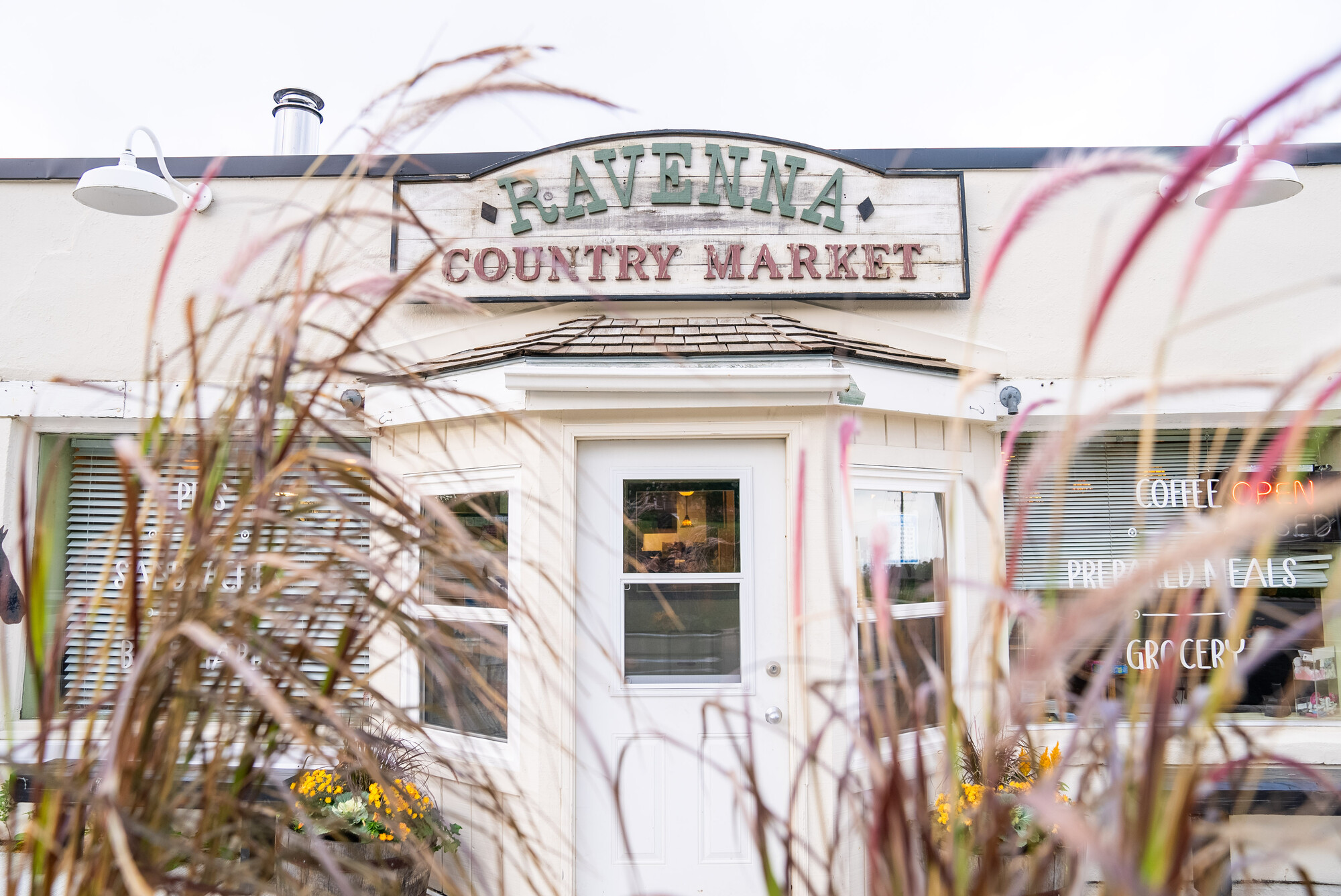 Exterior of Ravenna Country Market, a small shop with signage above the door, partially framed by tall ornamental grasses.