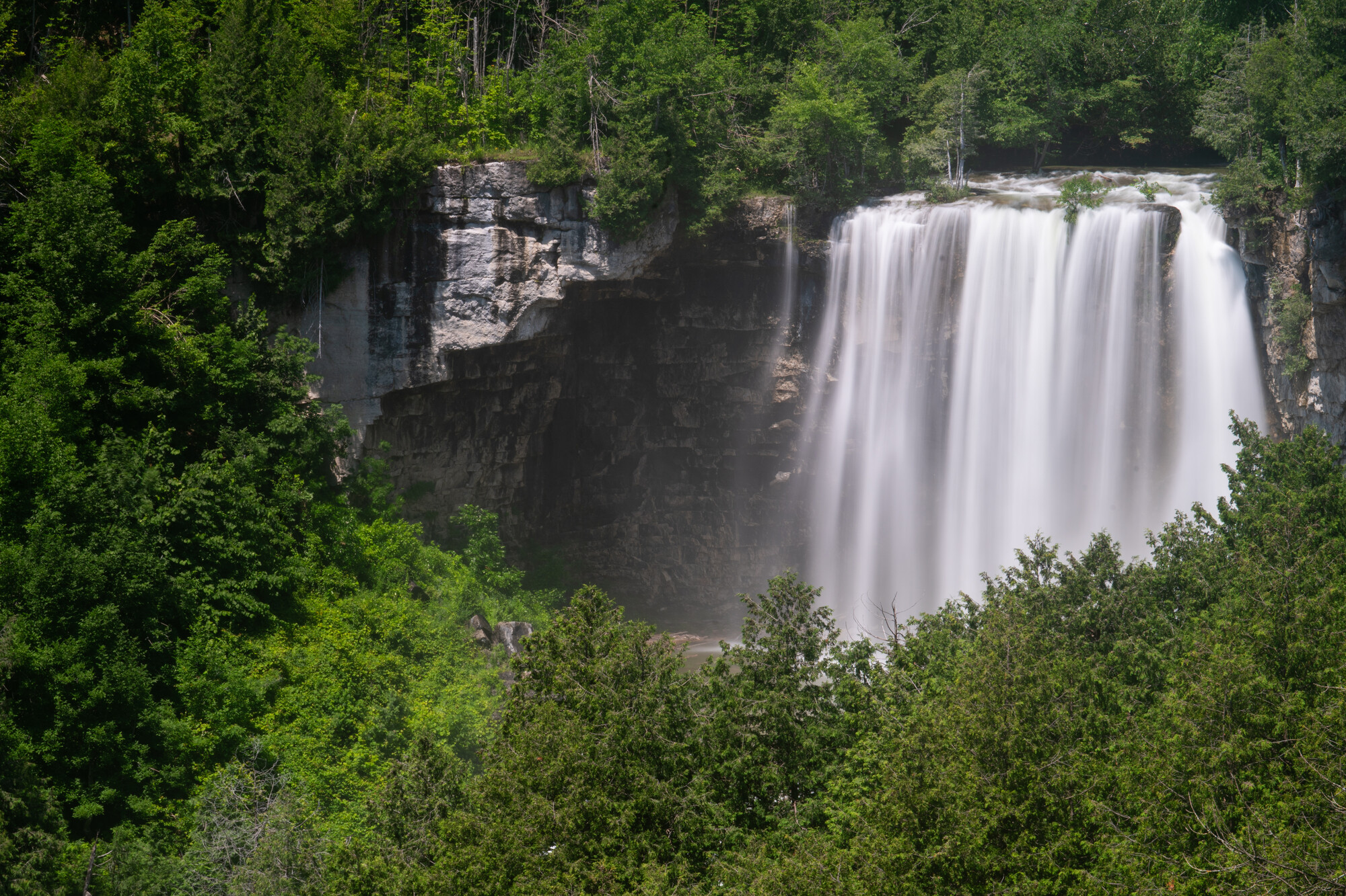 Wide waterfall cascading over a rocky cliff, surrounded by dense green forest on a bright summer day.