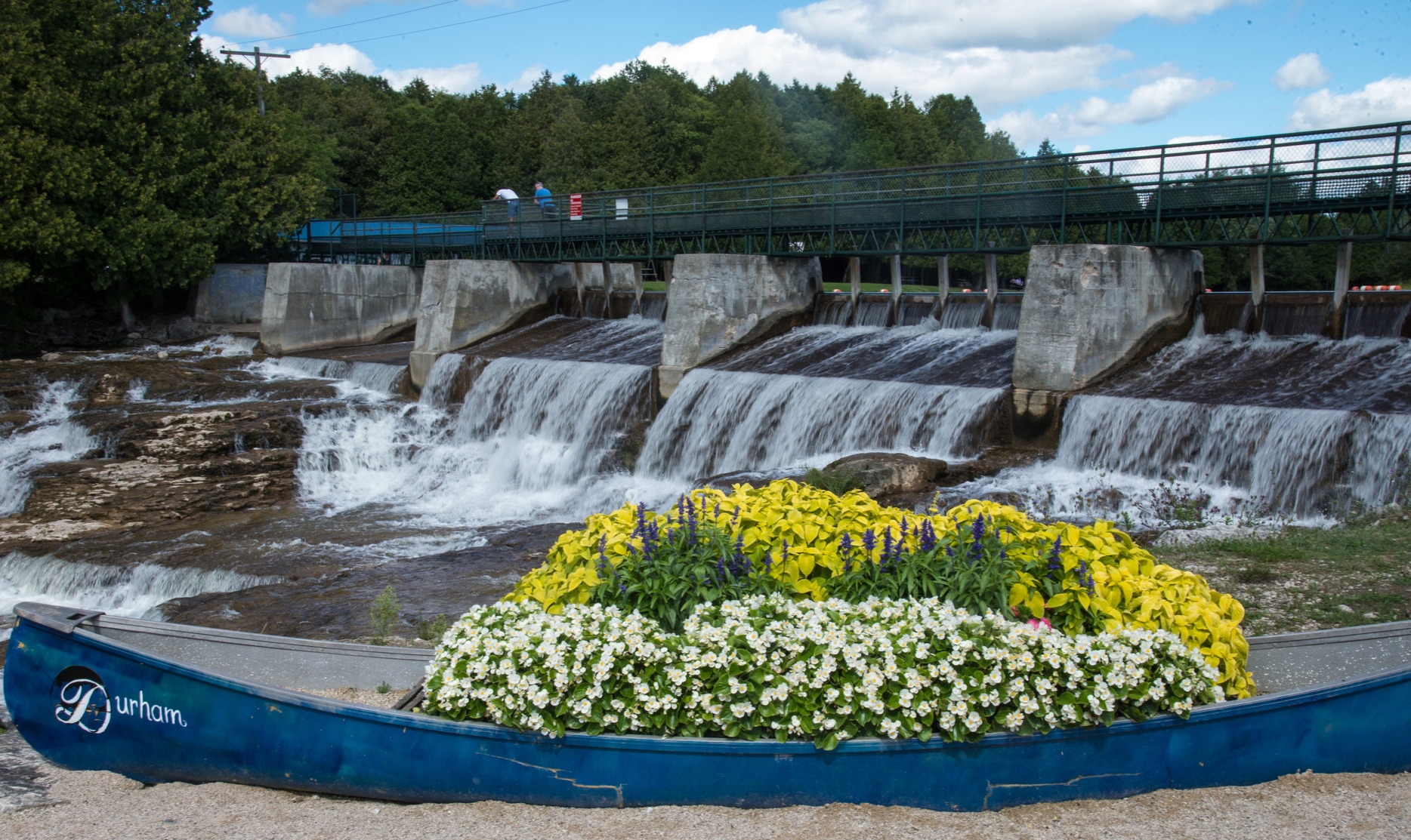 Water cascading over a dam behind a blue canoe filled with bright flowers.
