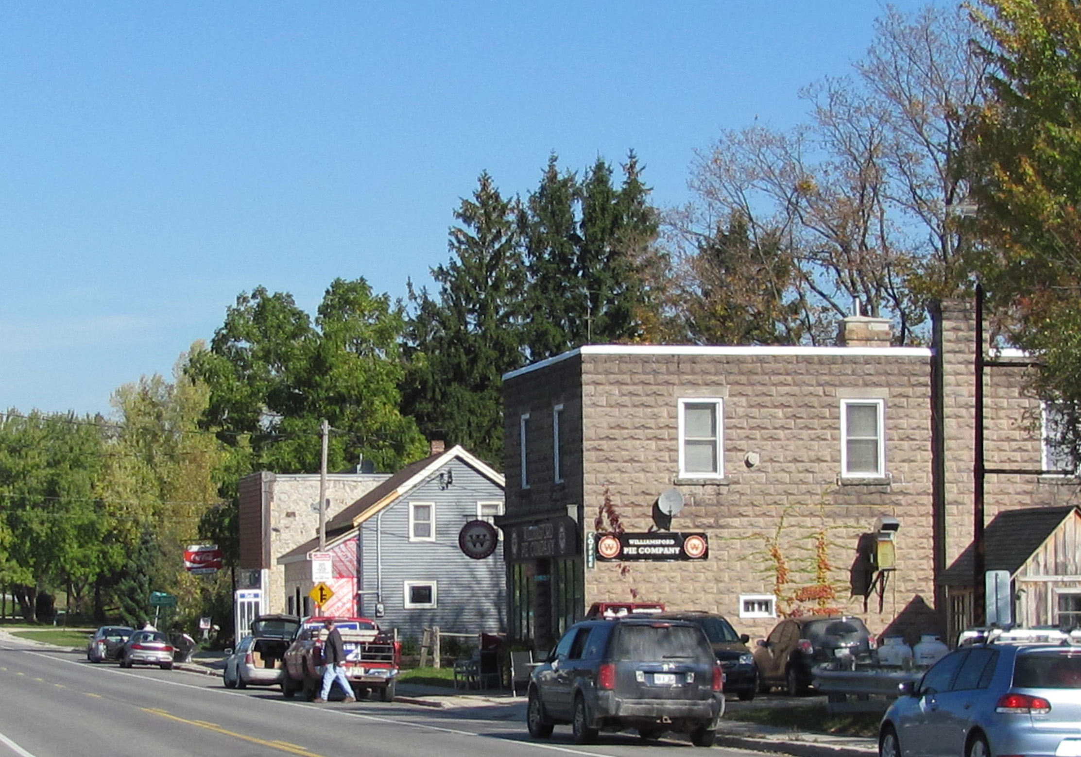 Small-town street with stone and wood buildings, parked cars, and trees in the background.