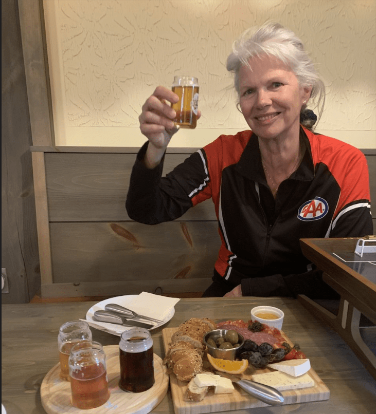 A smiling person raises a sampler glass of beer while seated at a table with food and drink laid out.