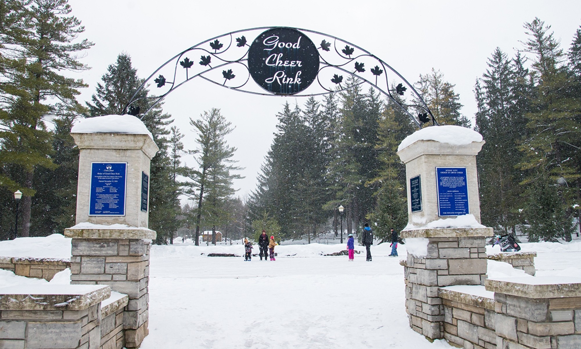 Stone columns hold up a sign declaring "Good Cheer Rink" in front of an outdoor ice rink.