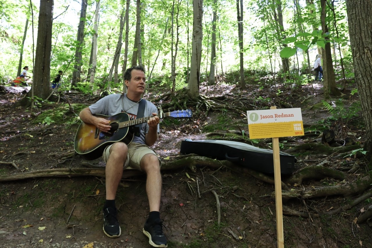 A person plays a guitar while seated in the woods.