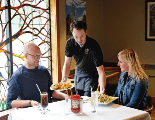 A server handing plates of food to two guests sitting at a table